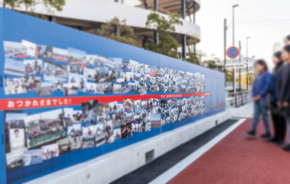 「橋梁完成記念パネルの展示風景」。Tamagawa Sky Bridgeの完成を記念し、工事関係者の写真と名前を掲載した大型パネルが屋外に設置され、見学者が鑑賞している様子。橋の建設に携わった多くの人々の努力を讃え、工事の進捗を時系列で整理し、写真と解説でわかりやすく伝えるデザイン。関係者への感謝のメッセージも記載され、訪れる人々に橋の完成までの歩みと貢献の大きさを実感してもらうための展示。